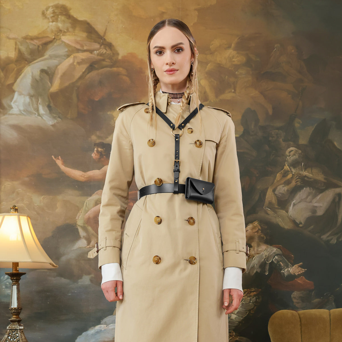 Woman in a beige trench coat standing in front of a classical, black leather harness with a belt and pouchpainting