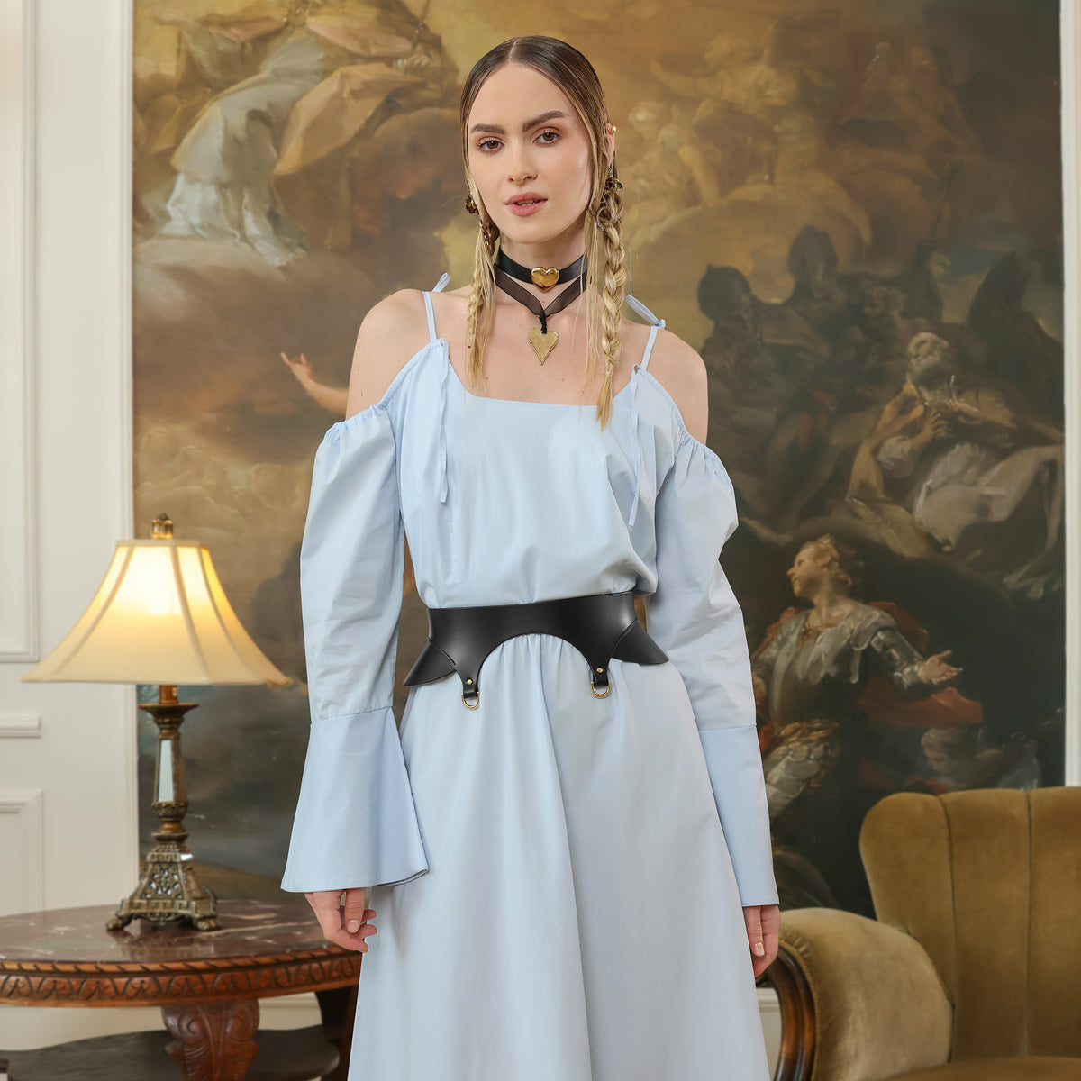 Woman in a light blue dress with a black belt standing in a room with classical painting in the background.