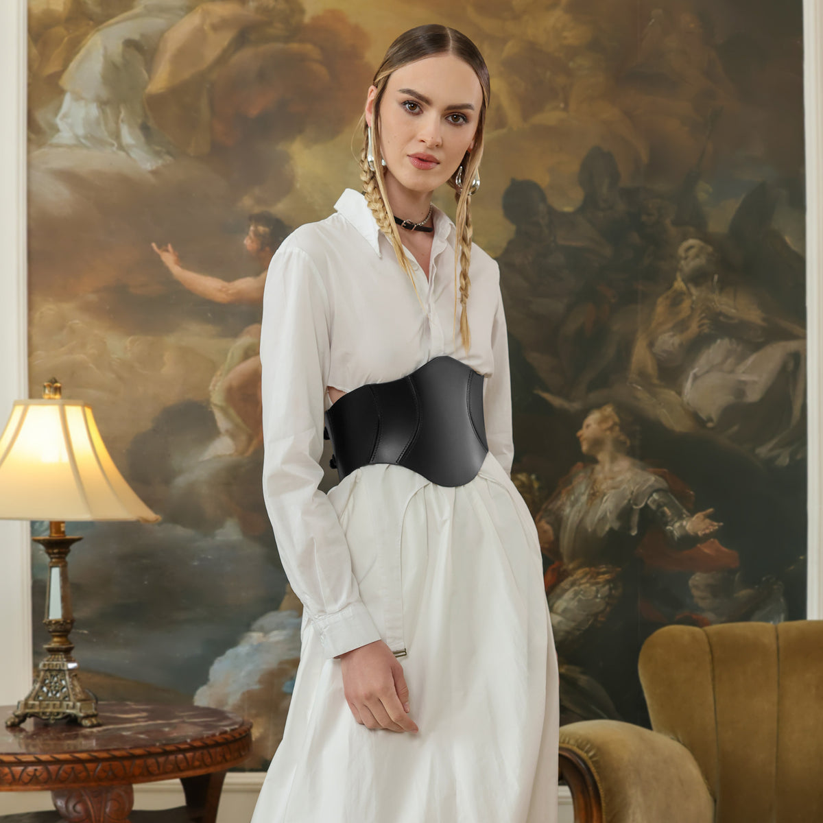 Woman in a white dress with a black corset standing in a room with classical painting and lamp.
