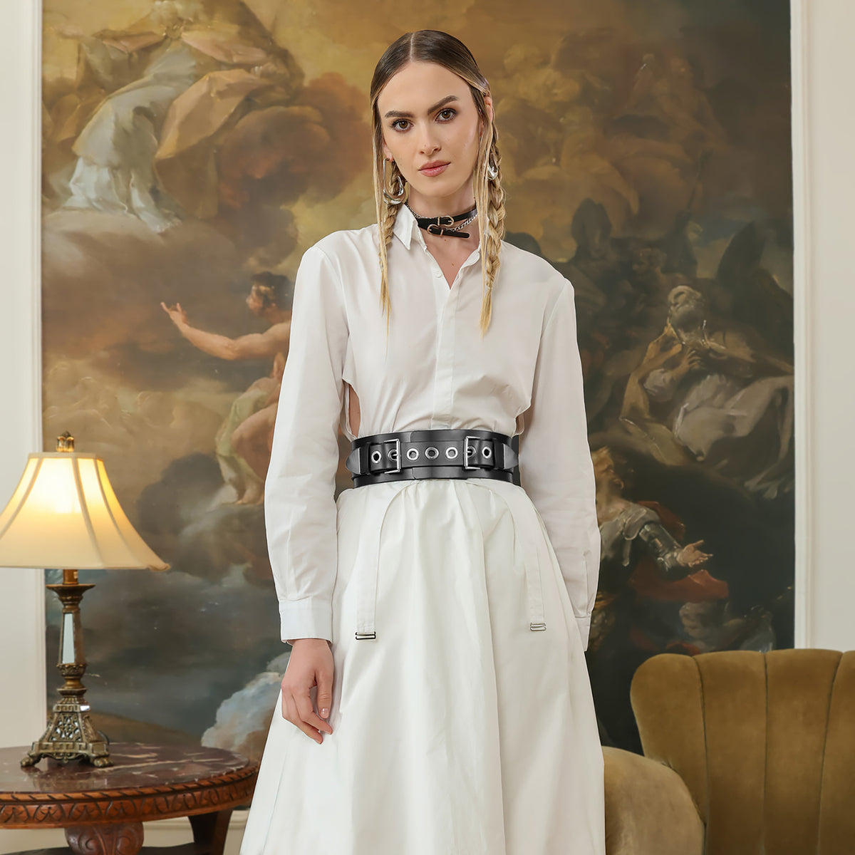 Woman in a white outfit with a black belt standing in a room with a large painting in the background.