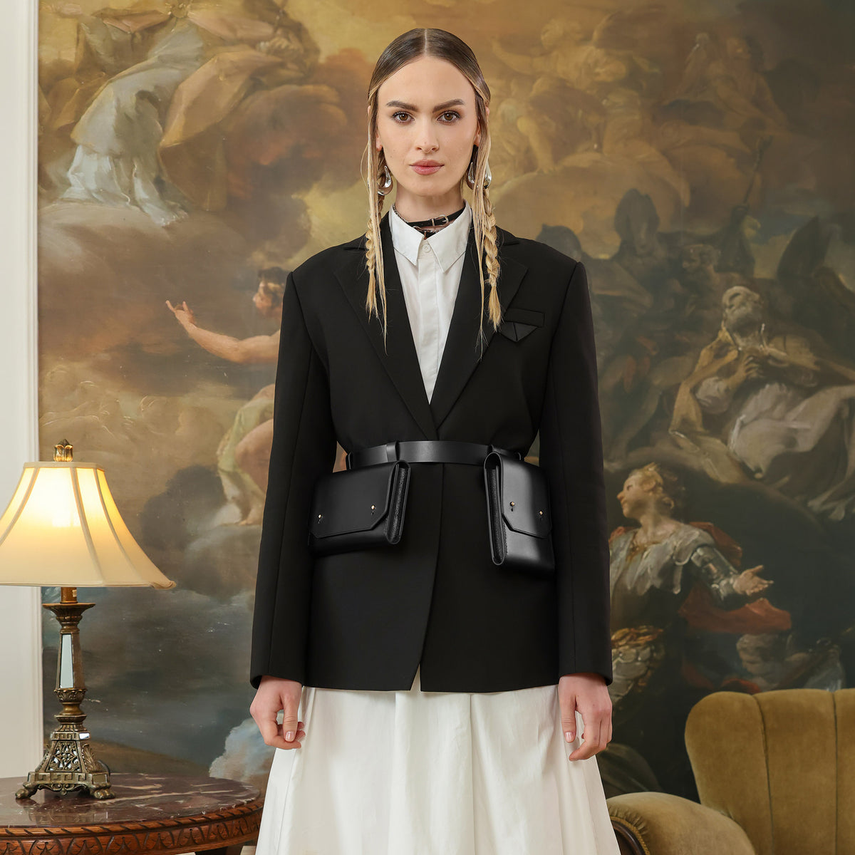 Woman in a black blazer with a belt bag standing in front of an ornate painting.