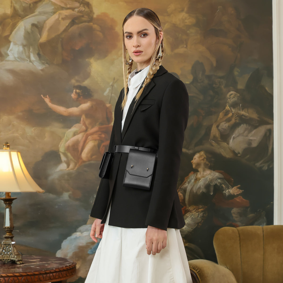 Woman in a black blazer and white dress standing in a room with classical painting on the wall.