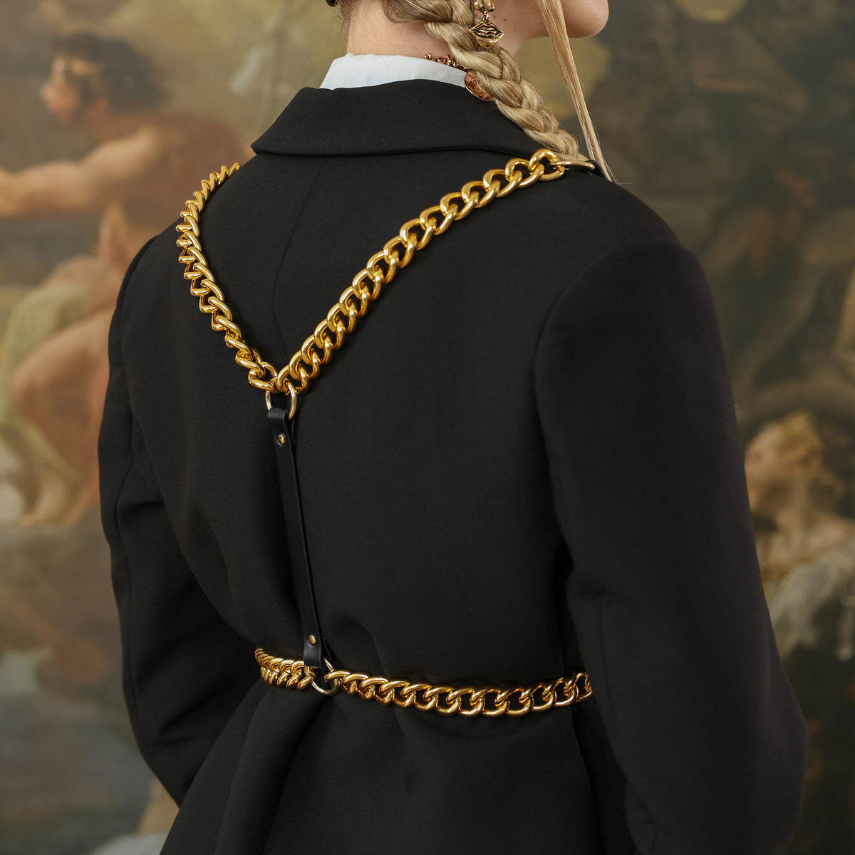 Person wearing a black coat with gold chains, standing in front of a classical painting.
