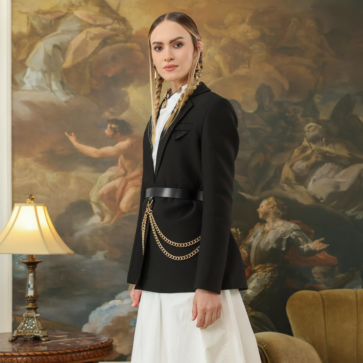 Woman in a black blazer with gold chains standing in front of a classical painting