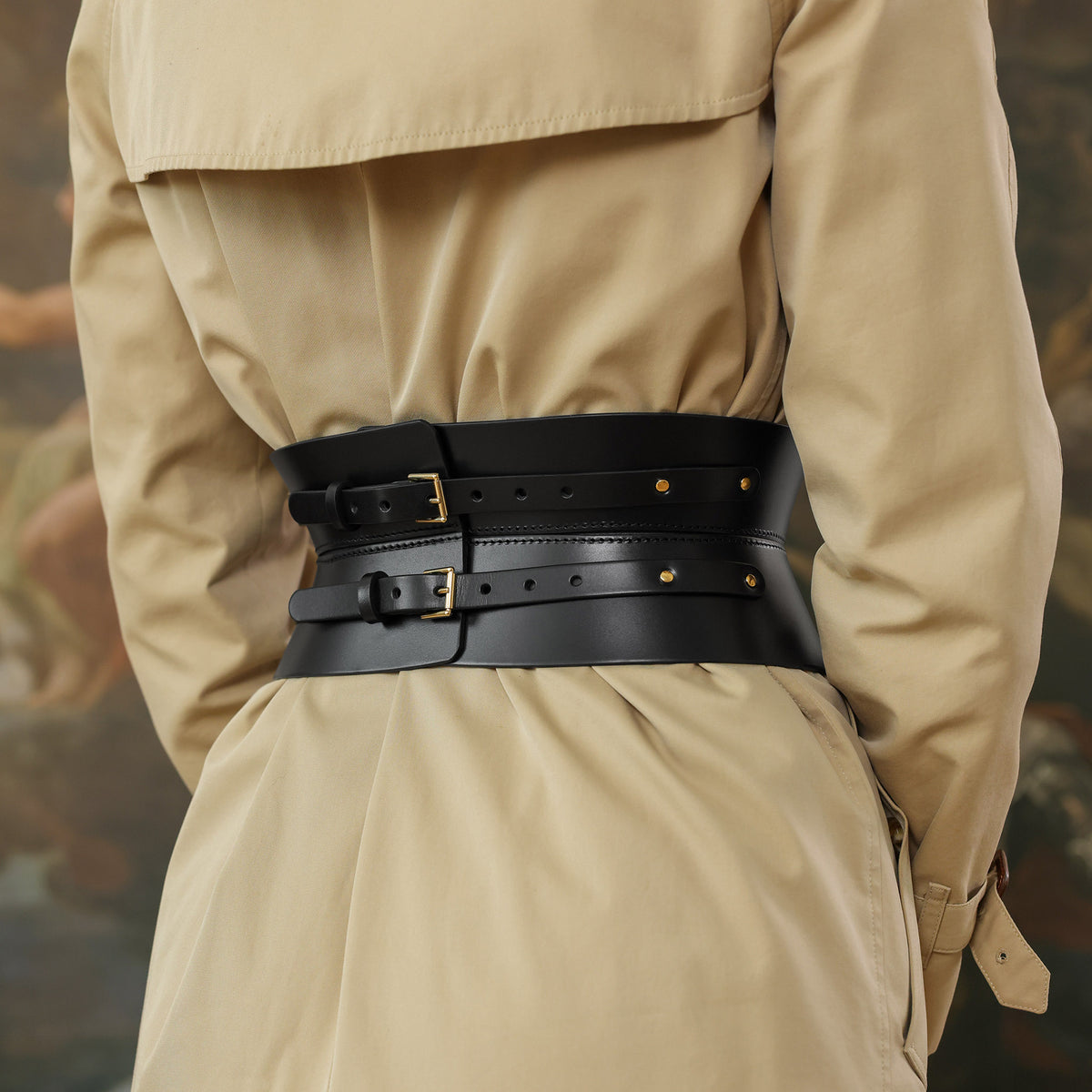 Person wearing a beige trench coat with a black belt featuring gold buckles.
