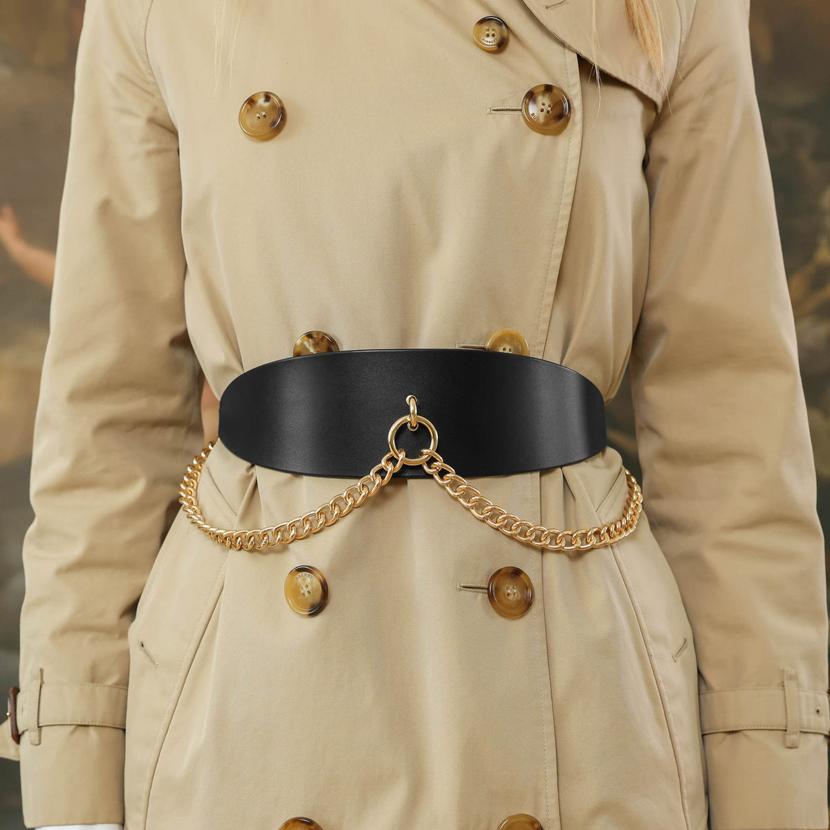 Person wearing a beige trench coat with a black belt and gold chain detail.