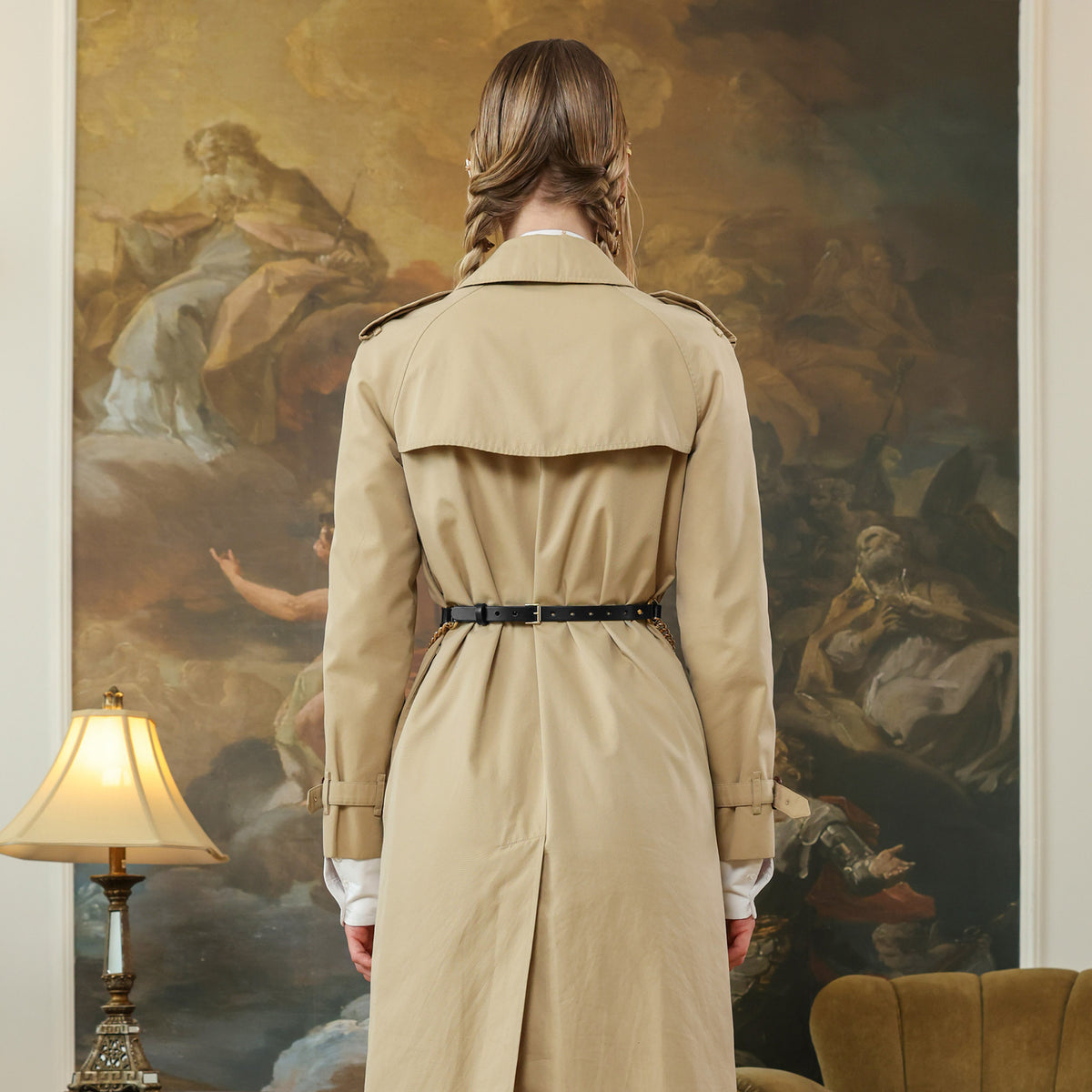 Woman in a beige trench coat standing in front of a classical painting. Black leather belt with gold chain