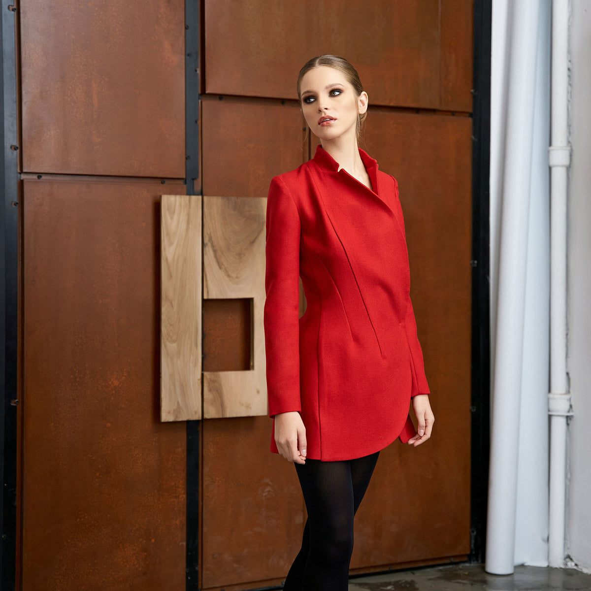 Woman in a red coat standing against a textured brown wall.
