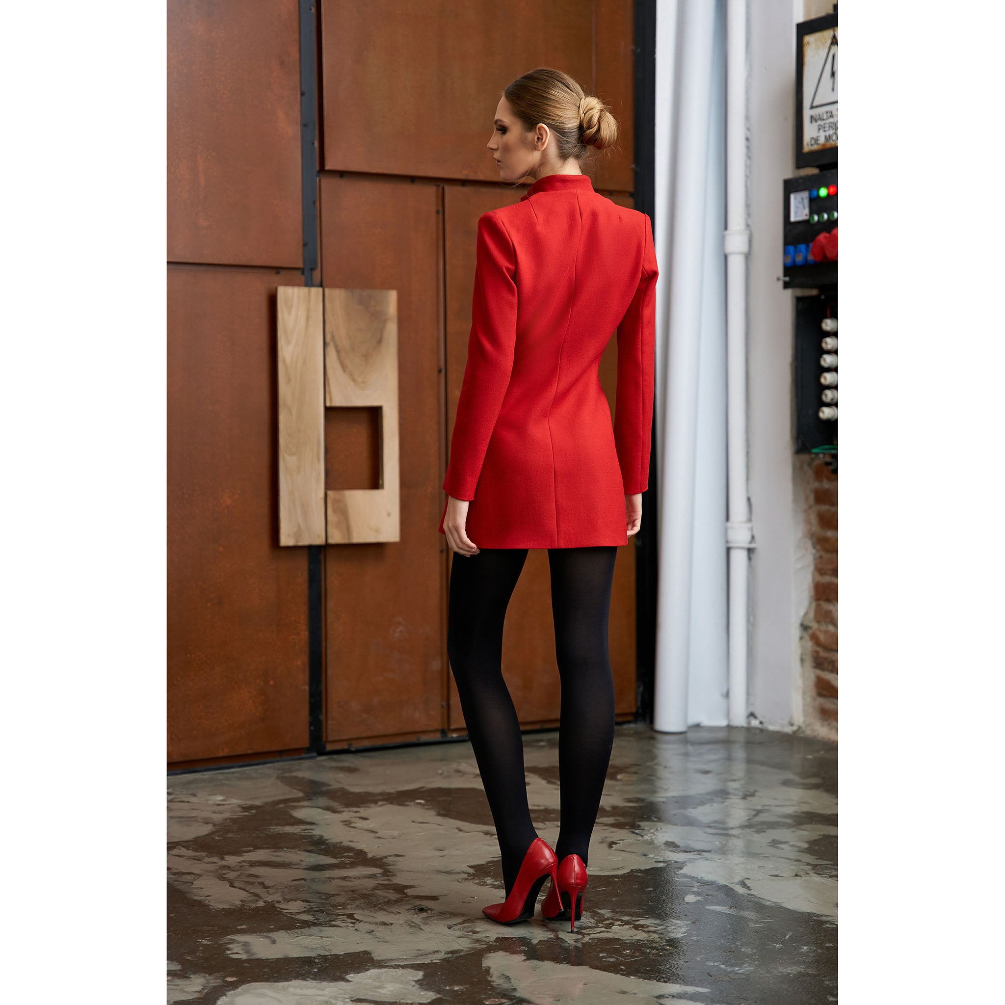 Woman in a red coat standing in a room with wooden panels and a white curtain.