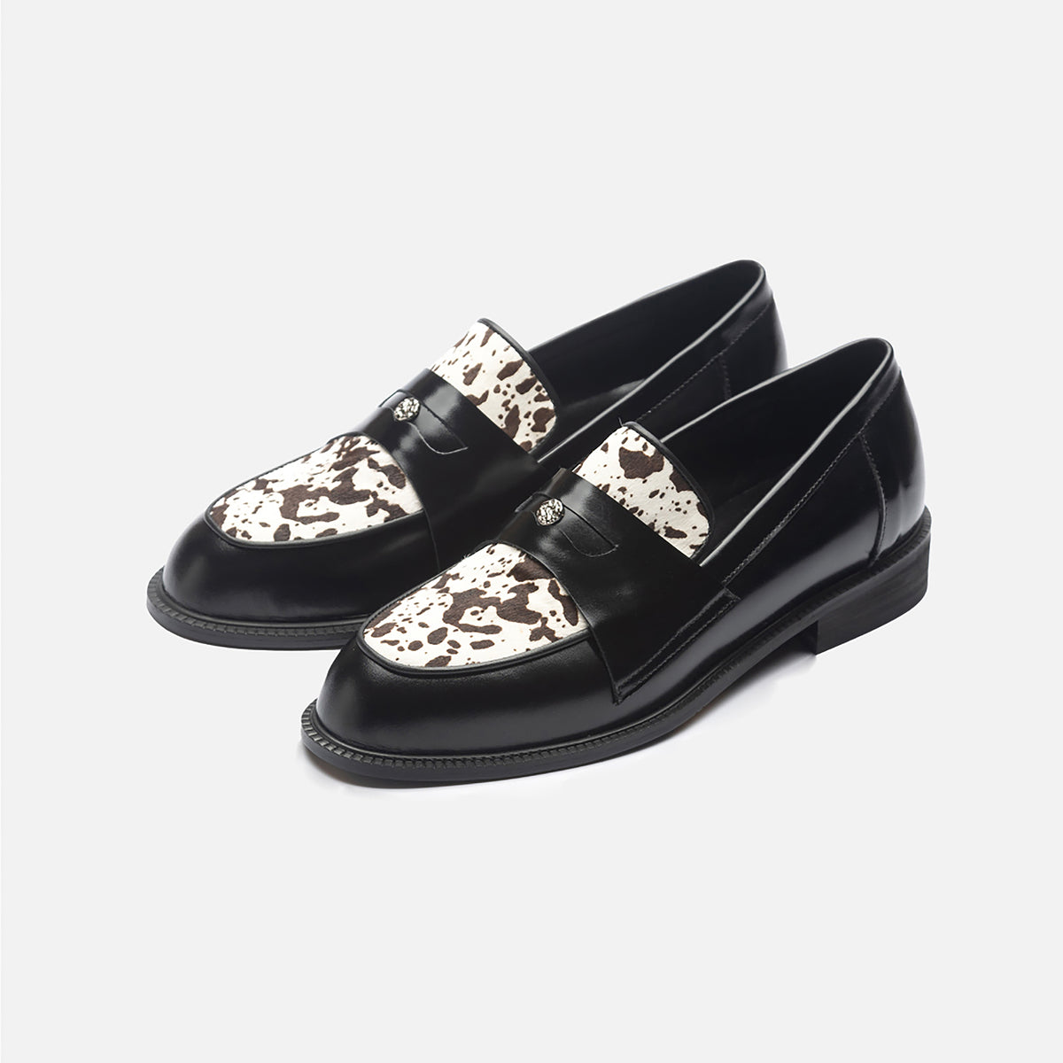 The THEN Black Leather Loafers with Cowhide Print Vamp feature a white and brown cowhide-print vamp, black straps, and are handmade in Korea. Displayed on a plain white background.