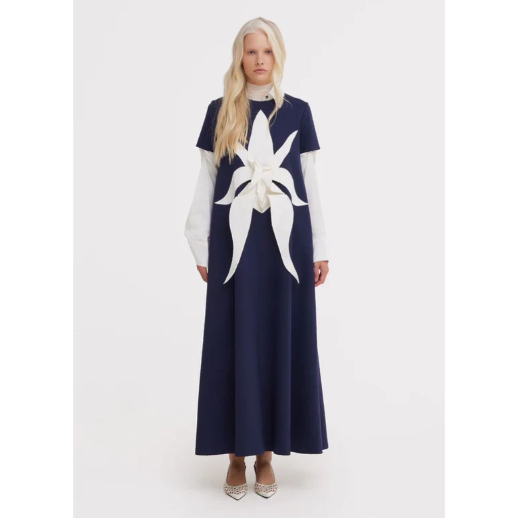 A woman with long blonde hair wears a JULIA ALLERT Ankle-Length Knit Dress With Flower Detail in dark blue, layered over a white long-sleeve top and pointed heels, standing against a plain white background.