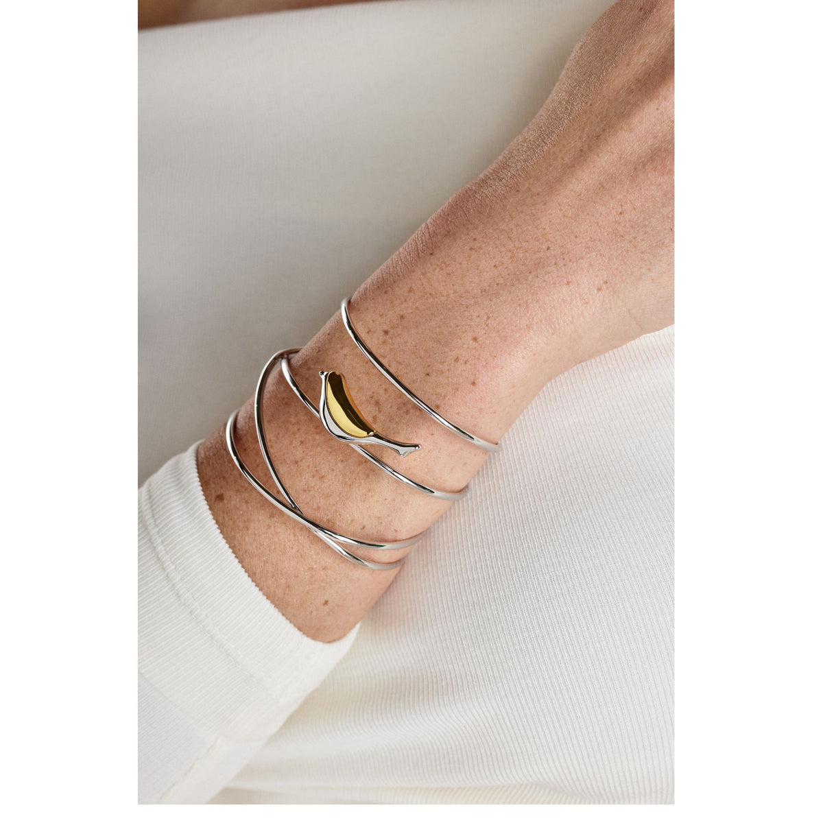 A close-up of a wrist wearing the ANNA PRATA Silver Aviva Bracelet from the Aviva Collection, featuring a hand-forged silver spiral and a yellow banana charm. The person is dressed in a long-sleeve white top.