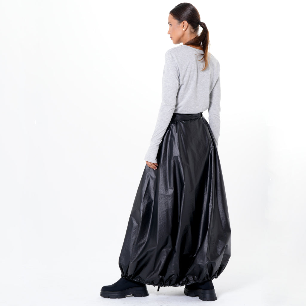 A woman with a ponytail stands facing away, wearing a long-sleeved gray top, METAMORPHOZA’s Voluminous Maxi Skirt with Adjustable Hem in Black, and black chunky boots against a plain white background.
