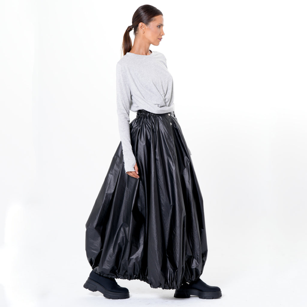 A woman stands sideways against a white background, wearing a light gray long-sleeve top and the METAMORPHOZA Voluminous Maxi Skirt with Adjustable Hem in Black, paired with chunky black platform shoes.