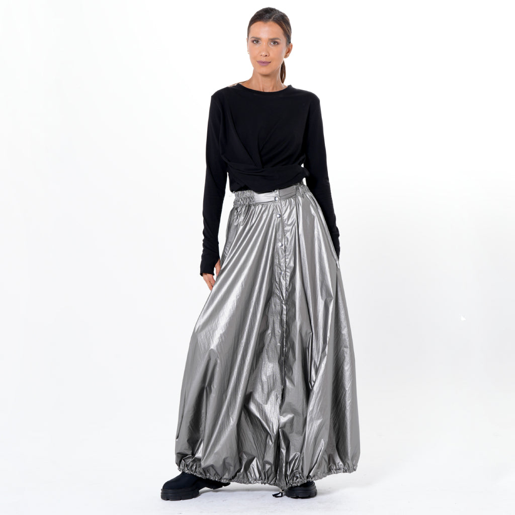 A woman stands against a white background wearing a long-sleeved black top, black shoes, and the METAMORPHOZA Voluminous Maxi Skirt with Adjustable Hem in Silver, gazing ahead with a neutral expression.