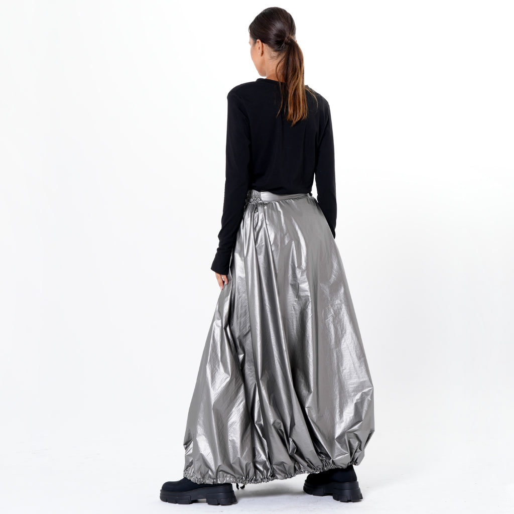 A woman with brown hair in a low ponytail wears a black long-sleeve top, chunky black boots, and the METAMORPHOZA Voluminous Maxi Skirt with Adjustable Hem in Silver, standing facing away against a white background.