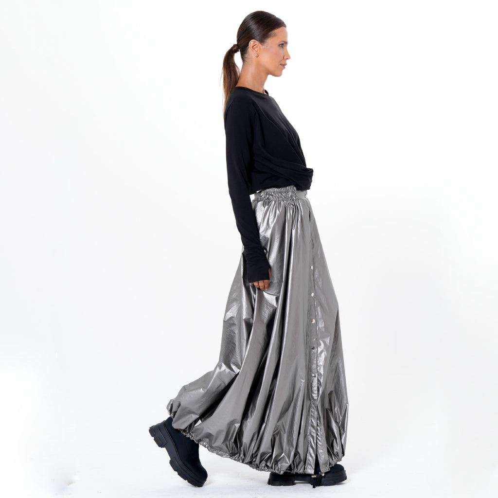 A woman with a ponytail wears a black long-sleeve top, black platform shoes, and the METAMORPHOZA Voluminous Maxi Skirt with Adjustable Hem in Silver as she walks to the right against a plain white background.