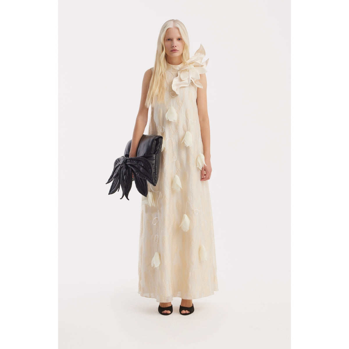 A woman with long blonde hair wears the JULIA ALLERT Ivory A-Line Flower Maxi Dress - Couture Gown, featuring fabric petals and a large bow at her shoulder. She accessorizes with a black bow-shaped clutch and black open-toe heels.