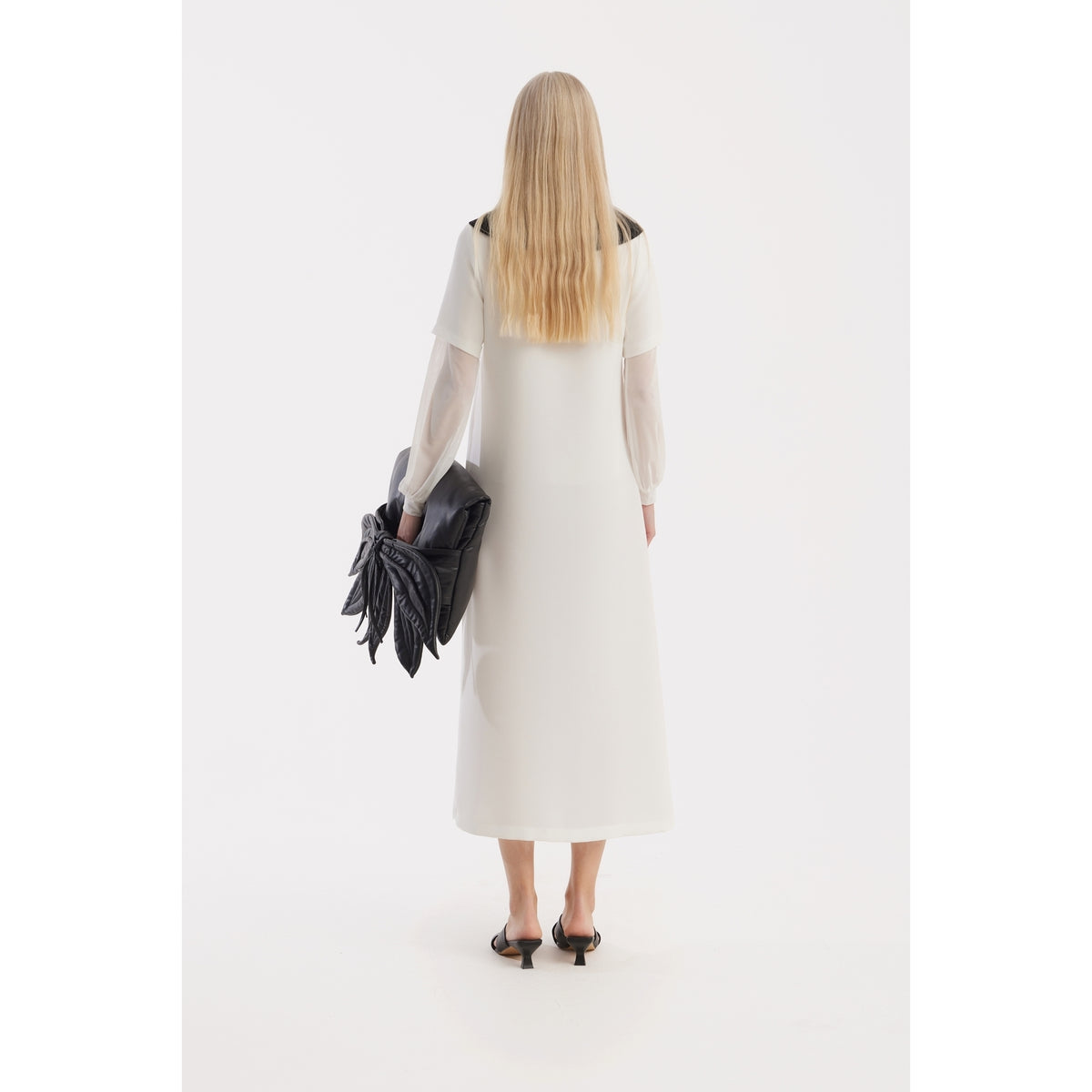 A woman with long blonde hair faces away, wearing the JULIA ALLERT Straight Midi Shirt Dress With Adjustable Collar - White. She pairs it with black sandals and holds a large black clutch against a plain white background.