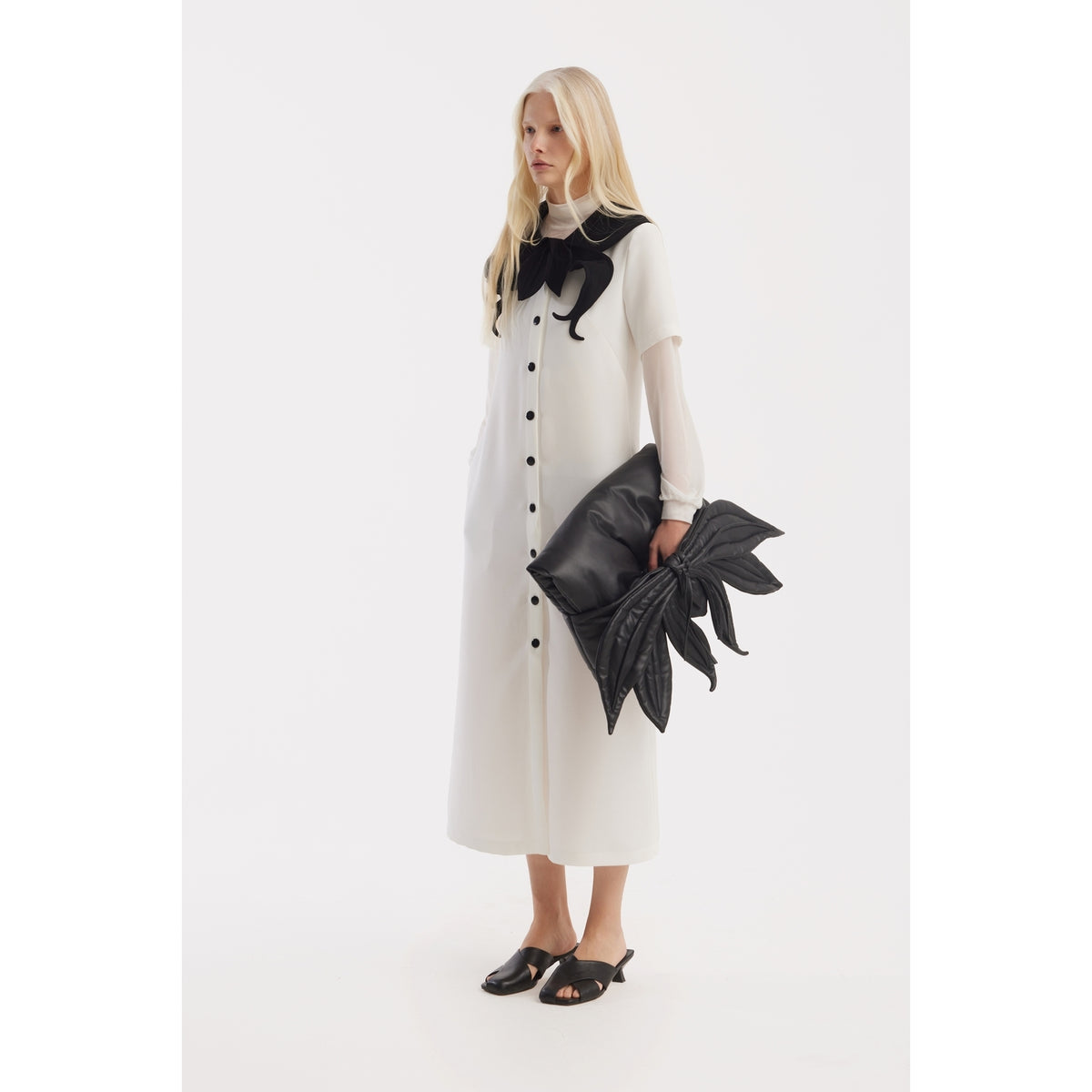A woman with long blonde hair wears a JULIA ALLERT Straight Midi Shirt Dress With Adjustable Collar in white, styled with an oversized black leaf-shaped clutch and black heeled sandals, standing against a plain white background.