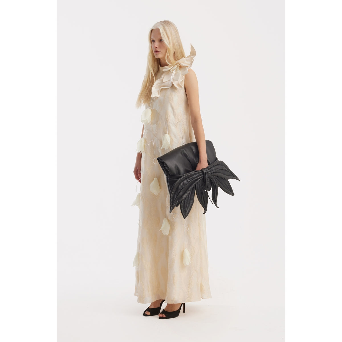 A woman with long blond hair wears the JULIA ALLERT Ivory A-Line Flower Maxi Dress - Couture Gown and black heels, holding an oversized black leaf-shaped clutch against a plain white background.
