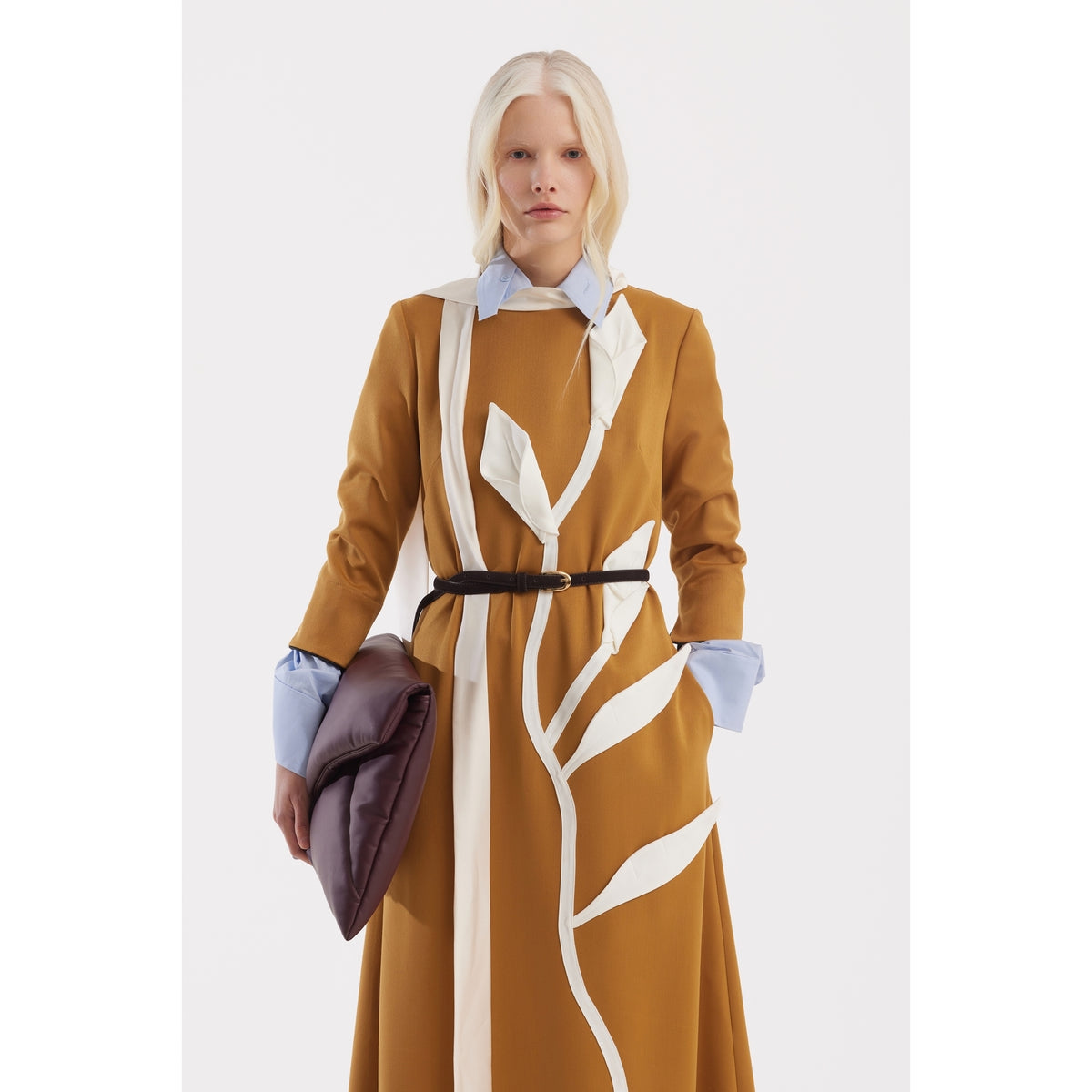 A person with long, light blonde hair wears the Art-Inspired Semi-Fitted Three-Quarter Sleeves Dress in mustard by JULIA ALLERT over a light blue collared shirt, holding a large dark brown clutch against a white background.