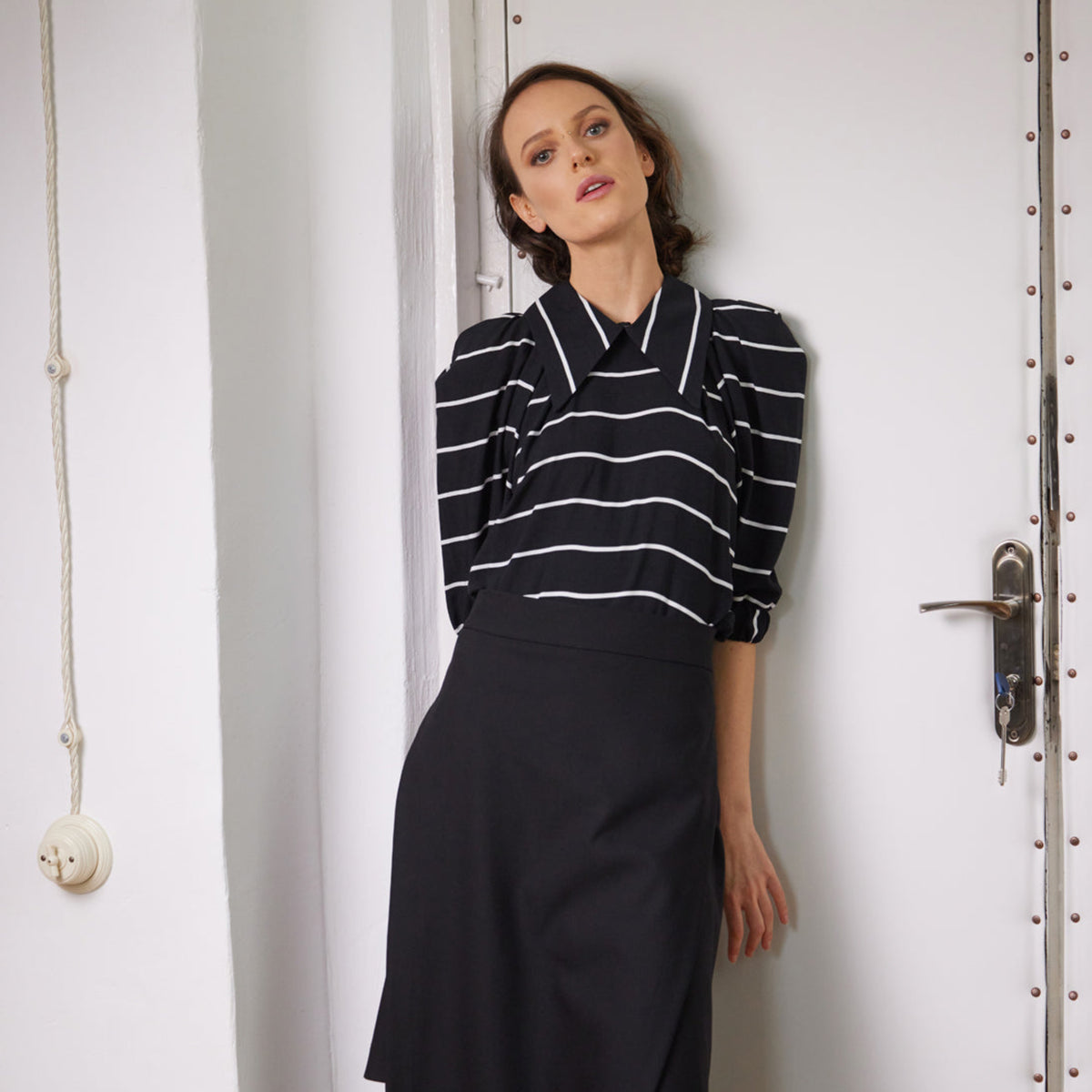 Woman wearing a black and white striped top and black skirt against a white wall.