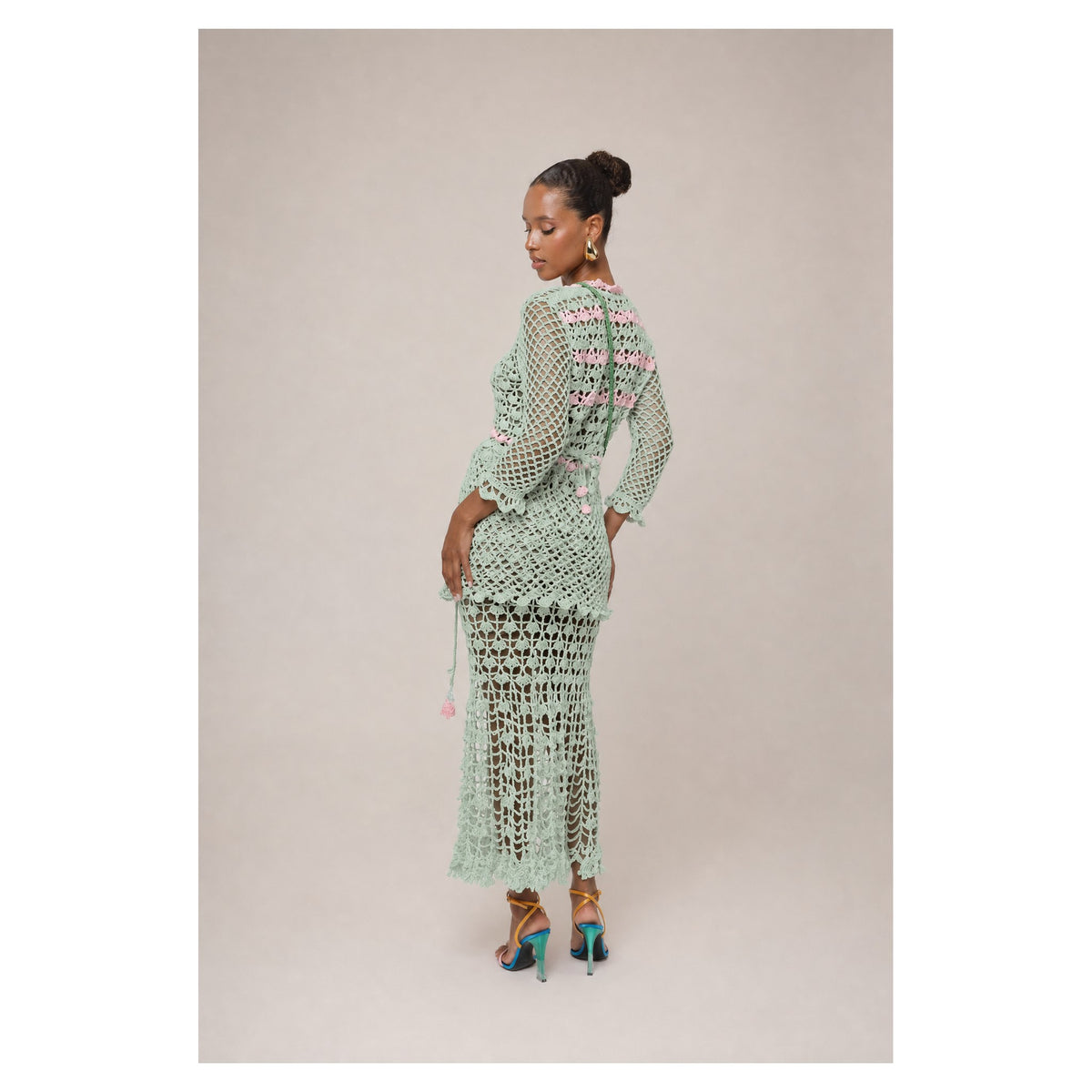 A woman poses in the California Mint Handmade Crochet Shirt by ANDREEVA, featuring light green crochet with pink accents. She pairs it with colorful heels and gold jewelry, her hair styled in a bun, against a simple beige background.