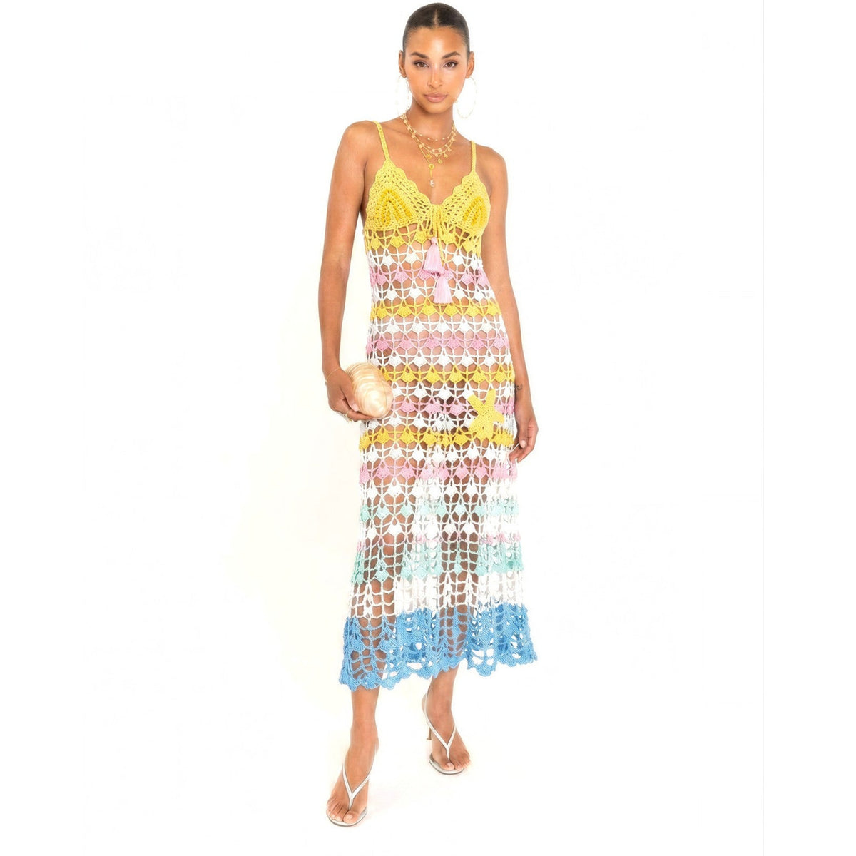 A woman poses in the ANDREEVA Flower Marine Handmade Maxi Crochet Dress, featuring floral patterns in yellow, pink, white, and blue. She accessorizes with a gold clutch, layered necklaces, strappy heeled sandals, and a sleek bun.