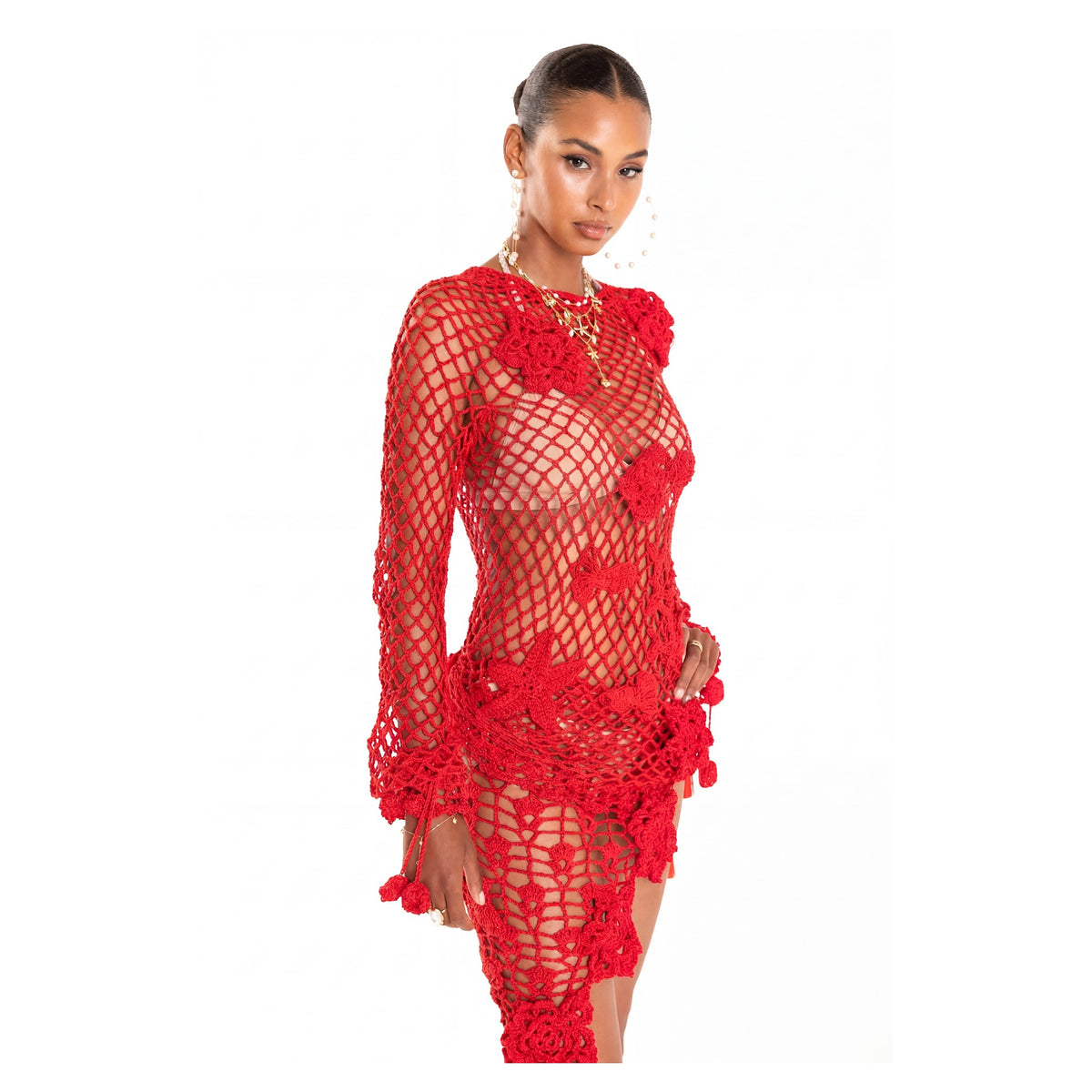 A woman models the Cherry Handmade Crochet Top by ANDREEVA, a sheer red cotton design with bold floral patterns, long sleeves, and a high neck. She styles it over a bikini with slicked-back hair and statement hoop earrings.