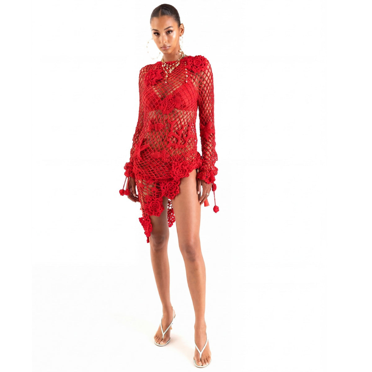 A woman models the ANDREEVA Cherry Handmade Crochet Cover-Up, a sheer red, long-sleeved dress with asymmetrical artisanal detailing, layered over a matching bikini and paired with white sandals against a plain white background.