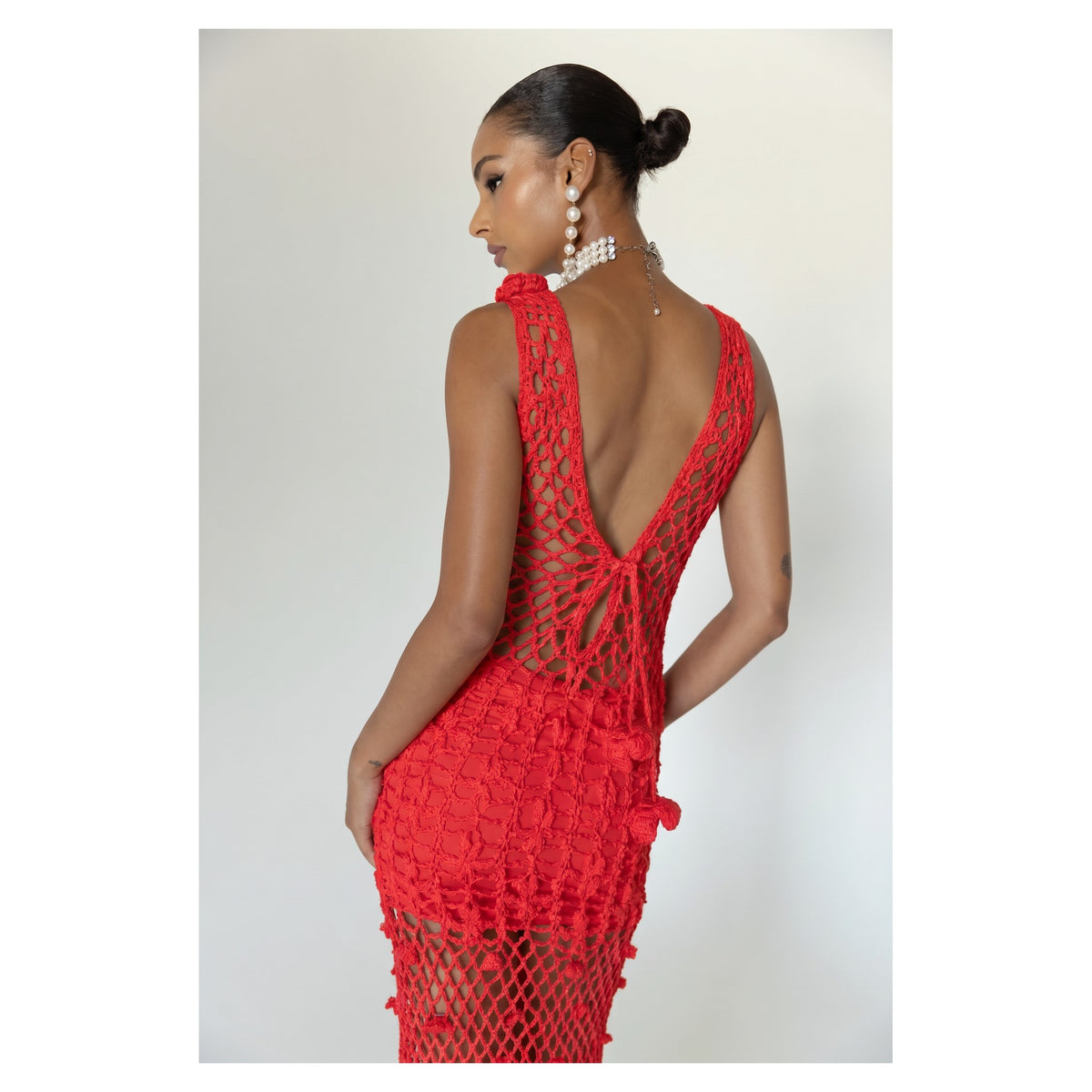 A woman poses in the Cherry Handmade Maxi Crochet Dress by ANDREEVA—a sleeveless, backless red crochet dress with a low-cut back. She wears pearl drop earrings and her hair in a sleek bun, standing against a plain light background.