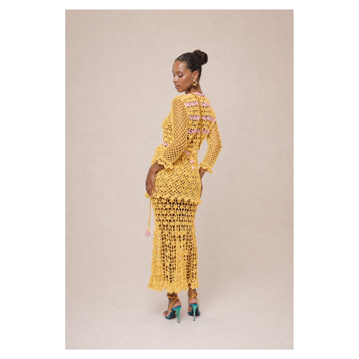 A woman wears the ANDREEVA California Poppy Yellow Handmade Knit Midi Skirt with a matching long-sleeved scalloped top and colorful heels, standing sideways against a beige background, looking over her shoulder with her hair up.